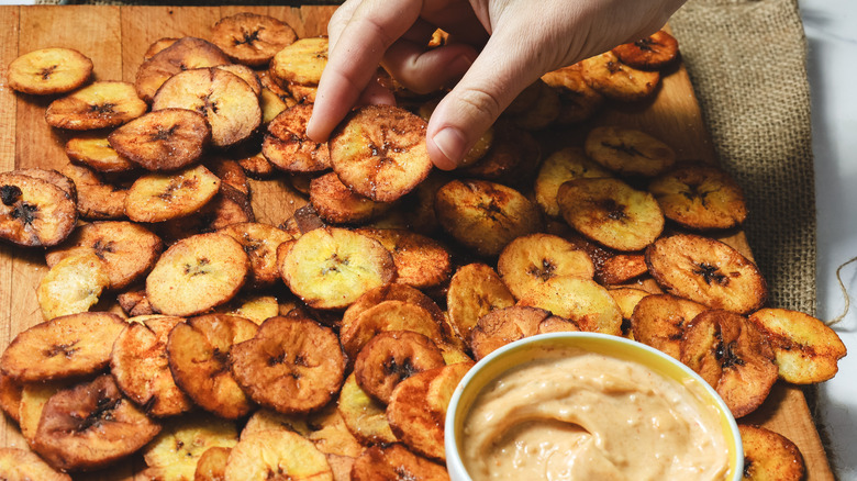 Hand holding a plantain chip over board with chips and sauce
