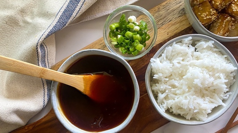 Bowl of sweet and sour sauce next to bowls of rice, crispy tofu, and chopped green onions