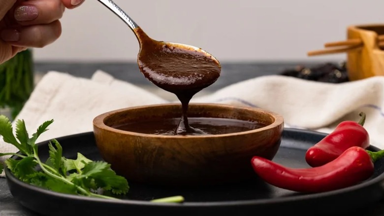Hoisin sauce in wooden bowl with spoon, next to sprig of cilantro and fresh red chile peppers