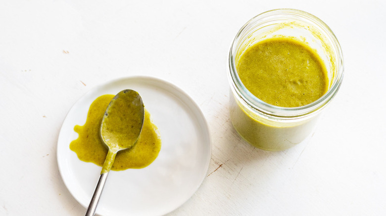 Jar of spicy green hot sauce next to plate with sauce-covered spoon resting on it