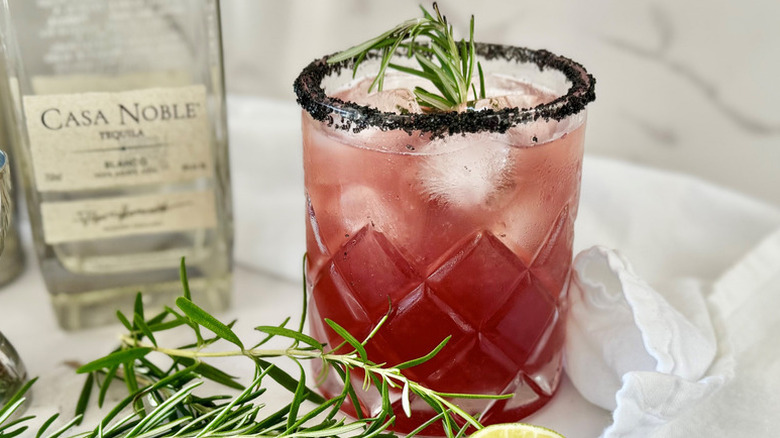 Pink cocktail in rocks glass with ice, rosemary, and a black salt rim