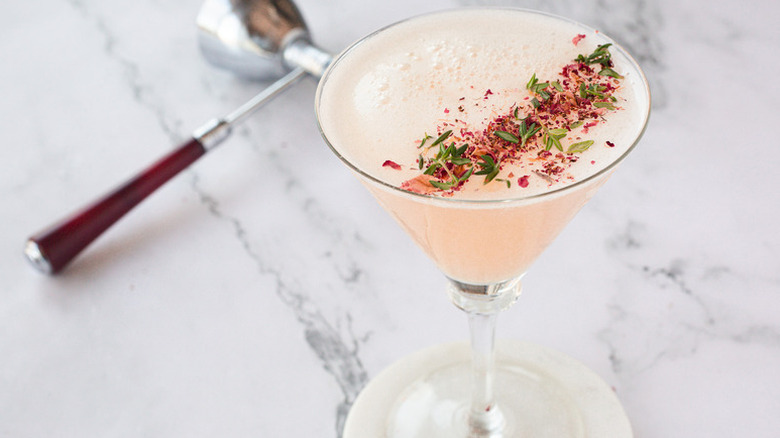 Blush pink cocktail in martini glass, garnished with crushed rose petals and thyme leaves