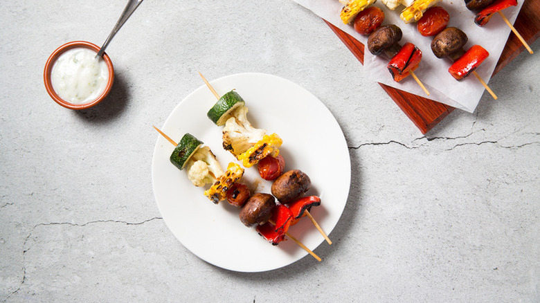 Grilled vegetable skewers on white plate next to small bowl of yogurt sauce