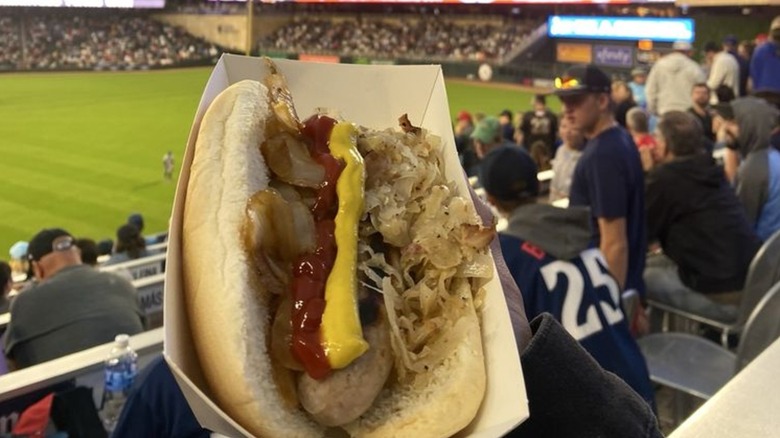Hand holding bratwurst with onions, sauerkraut, ketchup, and mustard in tray with blurred fans and field background