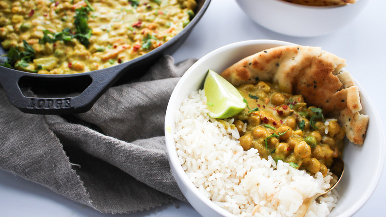 Chickpea curry in bowl with rice, naan bread, and lime wedge with the whole cast iron pan in the back