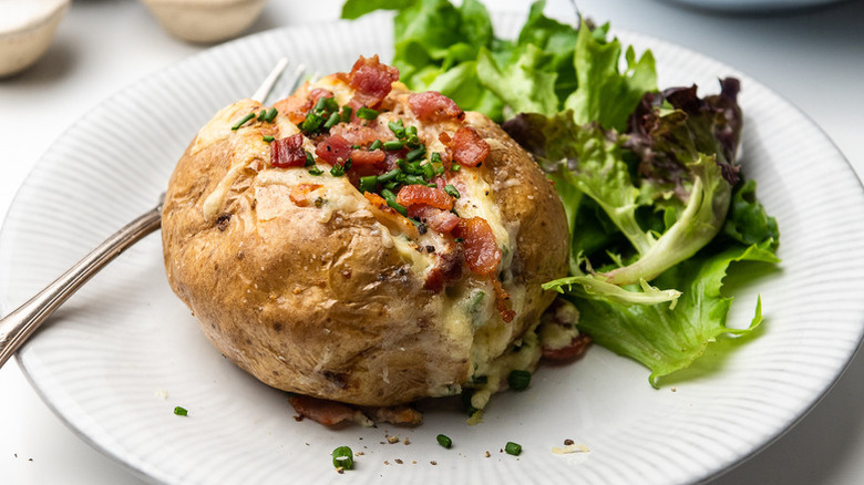 Baked potato stuffed with cheese, bacon and chives served on a white plate with fresh salad on the side