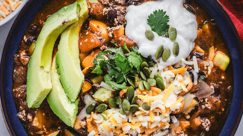 Close up of bowl of turkey and squash chili, topped with sliced avocado, cheese, sour cream, and pumpkin seeds