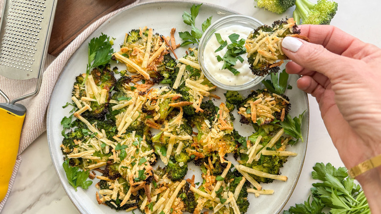 A person holding crispy, cheesy smashed broccoli and dipping it in a creamy dip over a whole plate of broccoli