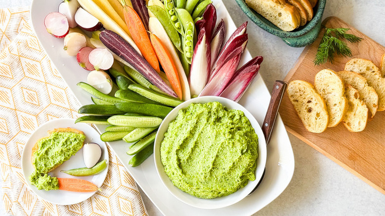 Bowl of creamy pea dip served with fresh sliced veggies and crostini