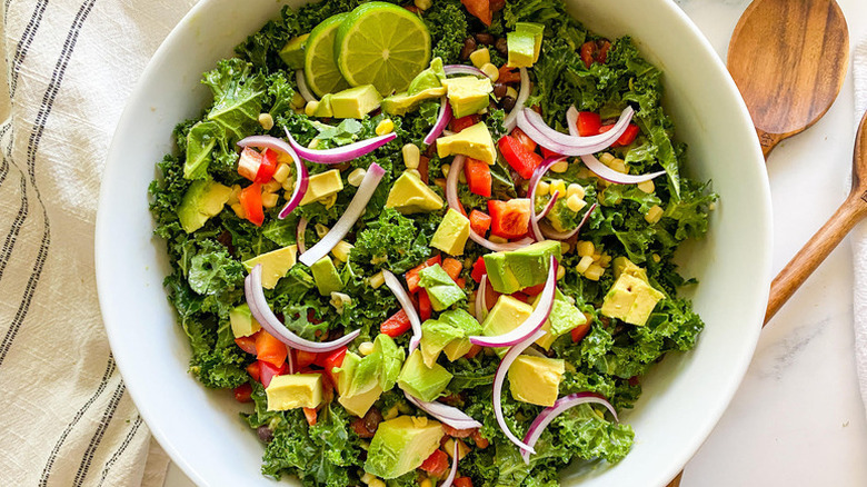 Top view of a white bowl of kale salad with avocado, red onion, and red bell pepper with wooden utensils on the side