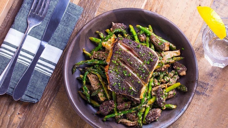 Bowl of pan-seared black cod with asparagus and morel mushrooms served on a dark plate with utensils and a glass on the side