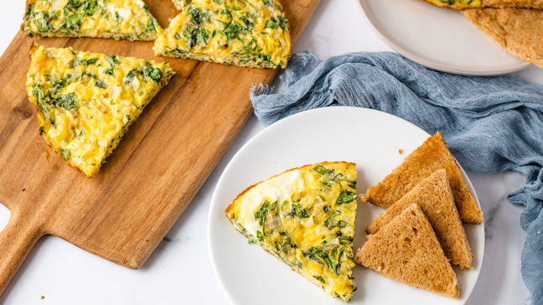 Slices of crustless spinach quiche on wooden board and plate with triangles of brown bread