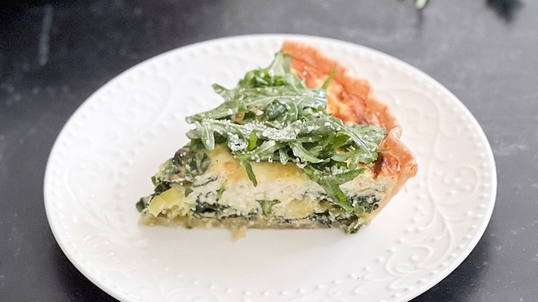 Slice of spring vegetable quiche in plate, topped with arugula