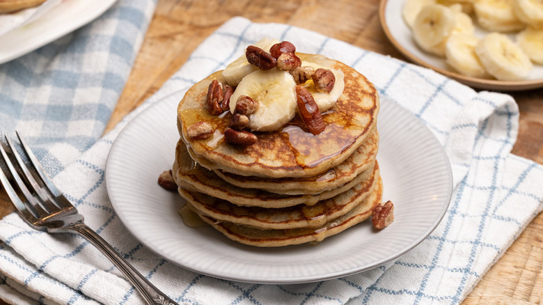 Stack of banana pancakes on plate, topped with chopped pecans and sliced banana
