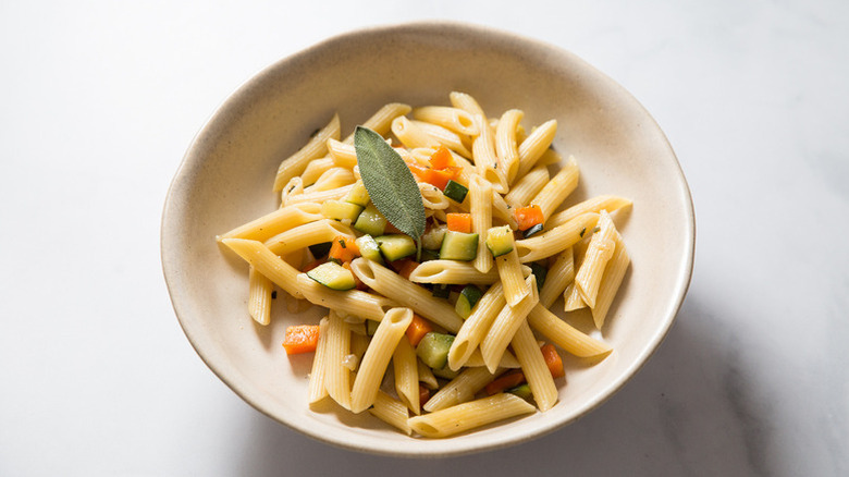 Bowl of penne pasta with diced vegetables and fresh sage.