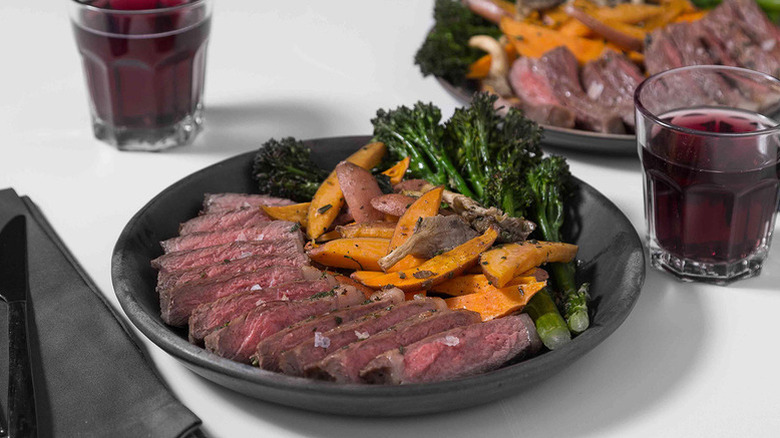 Sliced steak in dish with sweet potato wedges and broccolini aside a glass of red wine.
