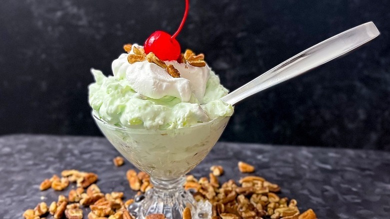 Watergate salad in a dessert glass, topped with Cool Whip, chopped pecans, and a maraschino cherry