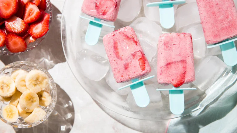 Strawberry Banana Popsicles on bed of ice next to bowls of cut fruit