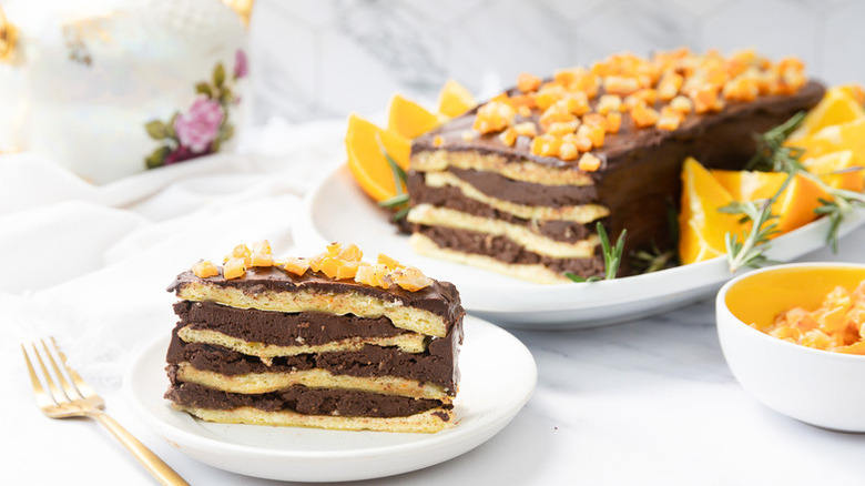Layered chocolate orange Passover cake slice served onto plate garnished with orange zest with the entire cake and oranges in the back