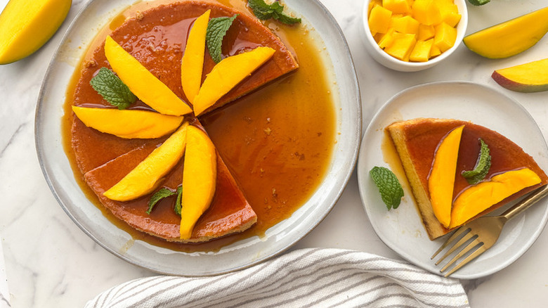 Top view of syrupy mango flan sliced and served onto plate with the whole flan and fresh mangoes around it
