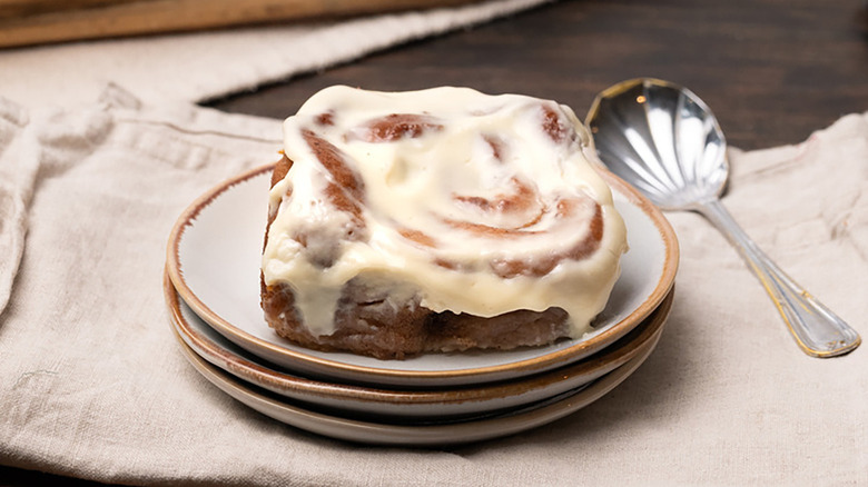 Frosting-topped cinnamon roll on plate with decorative dessert spoon on the side