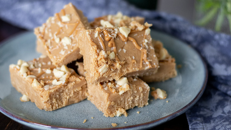Pile of peanut butter fudge on plate garnished with crushed peanuts and a peanut butter drizzle
