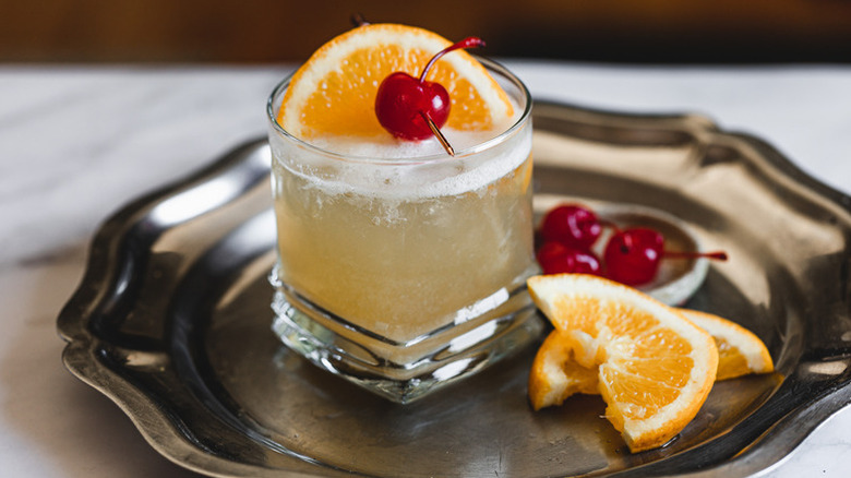 Frothy whiskey sour cocktail in glass, garnished with orange slice and maraschino cherry