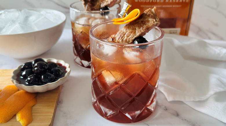 Manhattan cocktail in rocks glasses, each garnished with skewered steak, orange peel, and a luxardo cherry