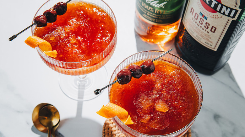 Frozen manhattan cocktails in coupe glasses, each garnished with skewered amarena cherries and an orange twist