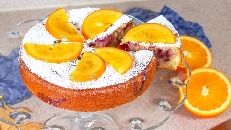 Sliced cranberry orange cake on glass cake stand, garnished with fresh orange slices