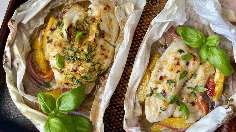 Baked tilapia and orange segments in unwrapped parchment paper, garnished with fresh basil