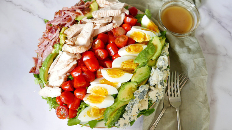 Colorful cobb salad on a serving plate next to forks and a bowl of dressing