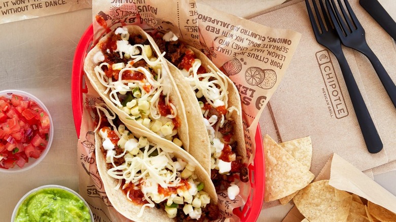 Red plastic basket holding three tacos with corn and cheese from Chipotle next to forks, napkins, and cups of salsa and guacamole