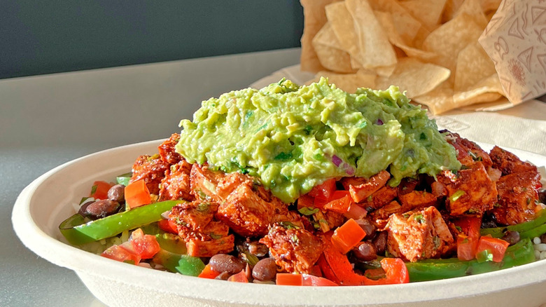Chipotle burrito bowl with chicken al pastor and guacamole on white table next to bag of chips and napkin