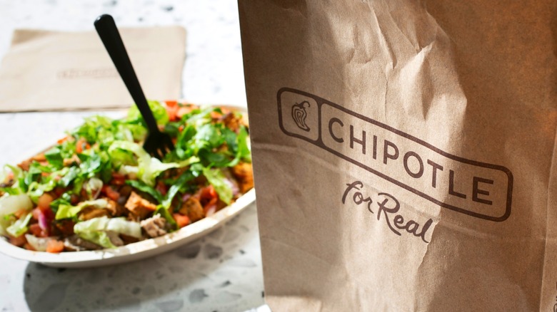 Brown paper bag with Chipotle logo and burrito bowl with lettuce and black plastic fork in background