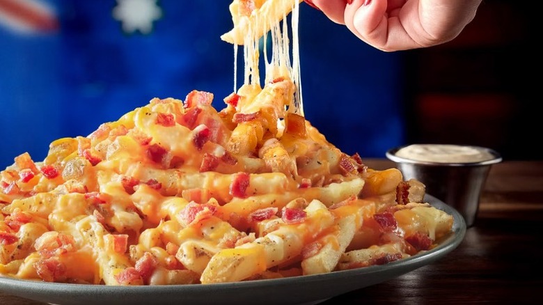 A closeup of a plate of Outback's Aussie Cheese Fries, with a hand pulling at fries with stringy cheese hanging off, and a side of ranch, on a wooden table