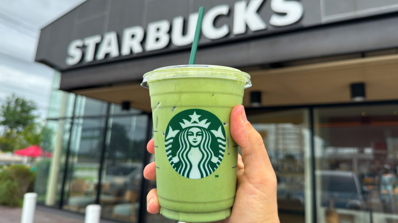 A hand holding an iced Starbucks Matcha Latte in front of the store