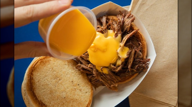A cup of cheese being poured onto a pot roast sandwich