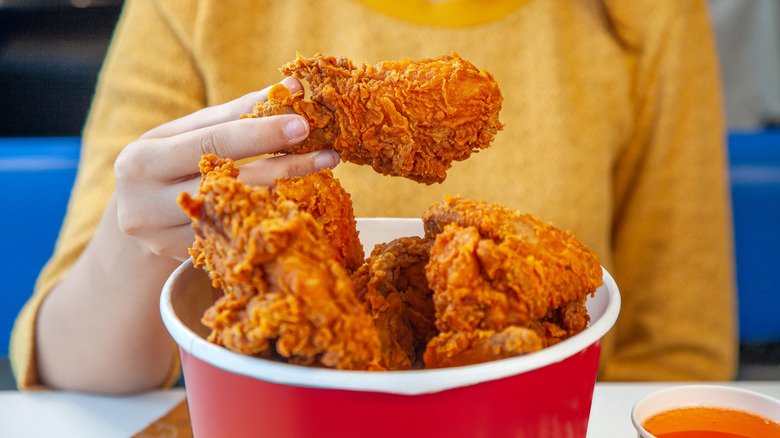 A person in a yellow shirt holding up a piece of fried chicken from a bucket