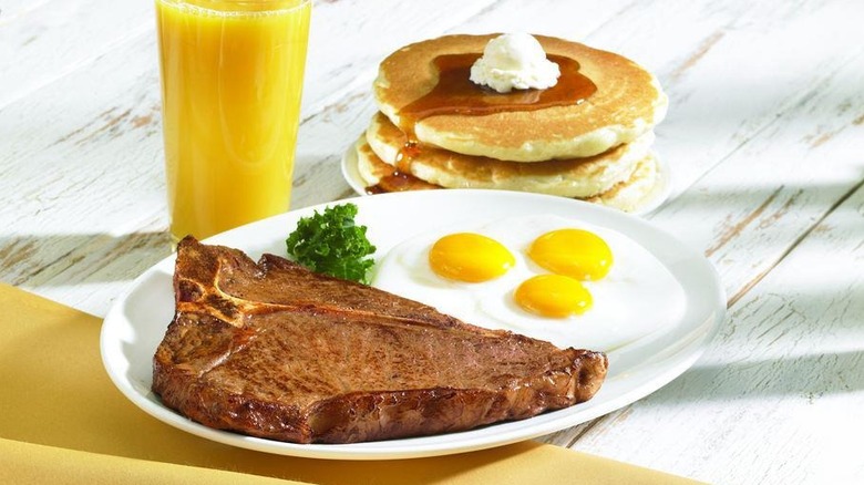 Plate of T-bone steak and sunny side up eggs next to glass of orange juice and stack of pancakes from IHOP
