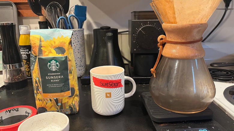 Starbucks Sunsera coffee bag, labeled coffee mug, and Chemex in front of coffee kettle and grinder on black counter