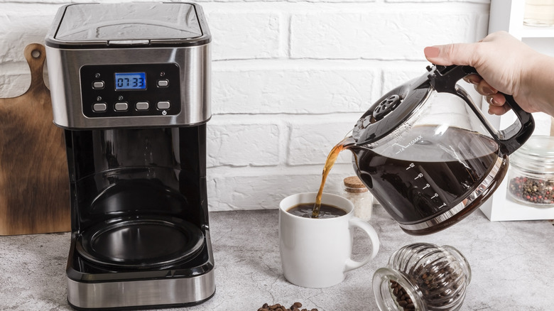 A close-up of a woman's hand pouring a cup of coffee from the carafe of stainless steel and black drip coffee maker on a kitchen counter