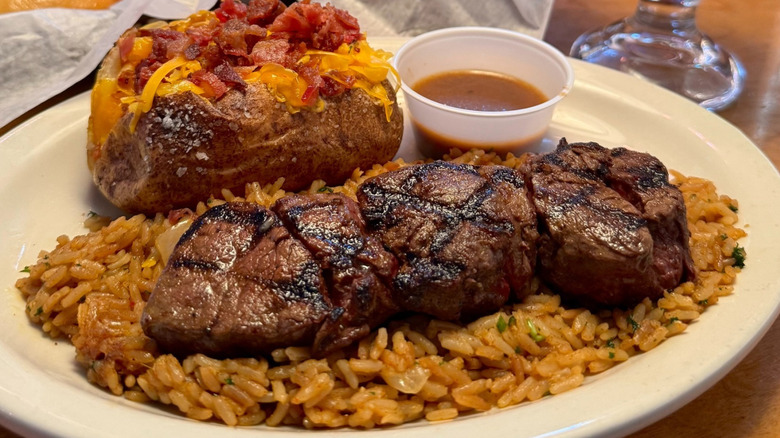 Plate of Texas Roadhouse filet medallions with rice, loaded baked potato, and cup of sauce