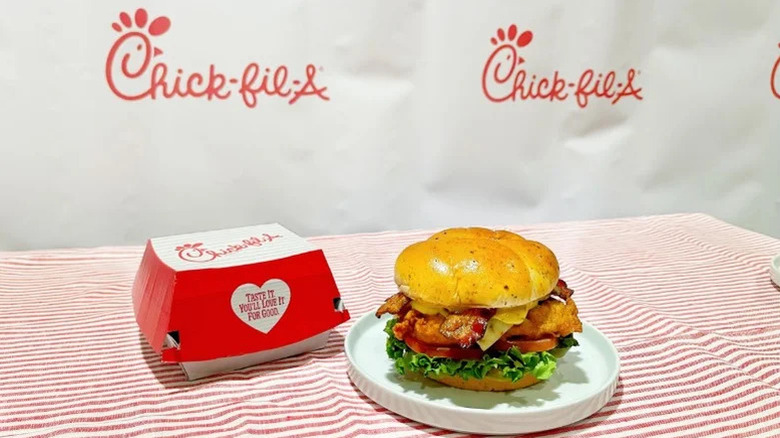 A Chick-fil-A sandwich sitting next to a Chick-fil-A takeout box on a red and white striped tablecloth with a Chick-fil-A backdrop