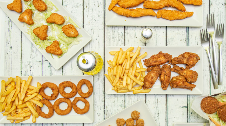 A variety of fried chicken dishes including nuggets, popcorn chicken, chicken strips, chicken wings served with onion rings and fries on a slat wooden table