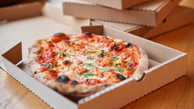 A whole pizza covered with cheese and sauce and some fresh herbs in a cardboard delivery box ,with other boxes stacked in the background