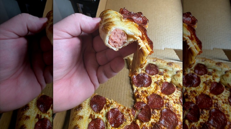 A hand holding a slice of hot dog stuffed crust pepperoni pizza. A bite has been taken out showing the hot dog inside, and the pizza itself is very limp