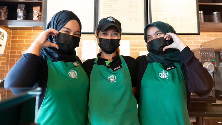 Three smiling female Starbucks barista in green aprons, wearing black face masks