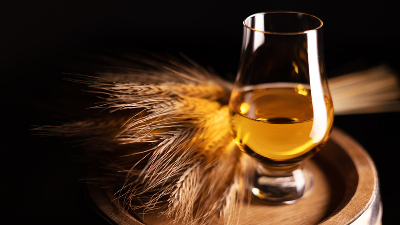 A glass of whiskey next to a bunch of rye on to of a barrel.
