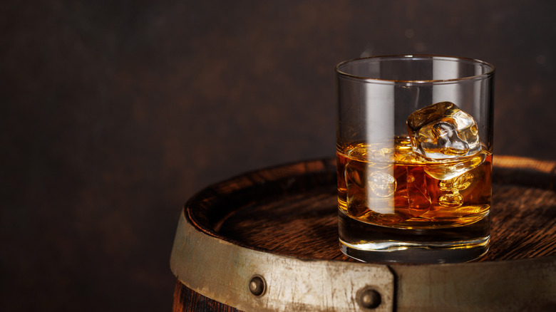 A glass of whiskey and ice on top of a wooden barrel.